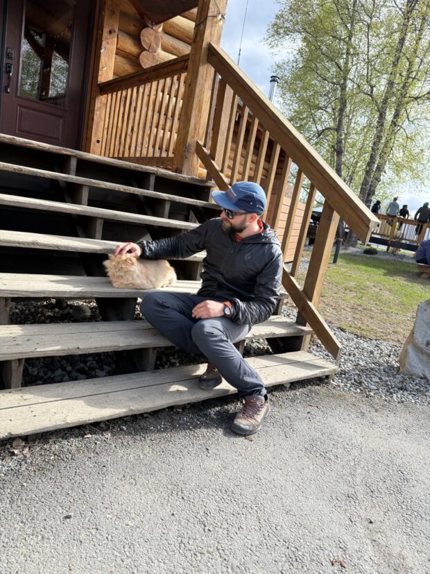 IMG Guide, Brad Swanson, sits on the front steps of a log building petting a very content orange cat