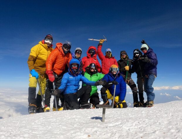 A team of climbers lead by Forrest Baldwin pose for a group photo on the top of Denali/McKinley in 2022 with the summit marker in the foreground.